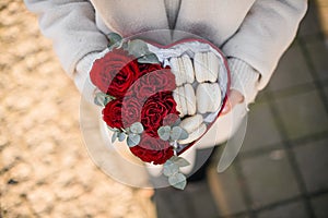 Woman holding heart shaped box with fresh red roses bugs and french sweets macarons in hands. Top view