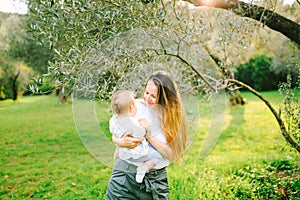 Woman holding her baby girl in her arms under an olive tree on a sunny day