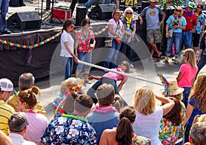 Woman holding her shoes and bending over backwards to dance under a limbo bar surrounded by people in tropicl clothing at party in