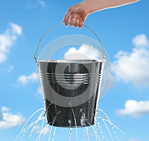 Woman holding leaky bucket with water against sky, closeup photo