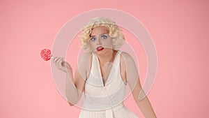 Woman holding a lollipop on a stick and shrugging her shoulders. Woman looking like Marilyn Monroe in studio on pink