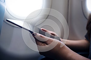 Woman holding and looking at black tablet pc next to an airplane window with clouds and sky background