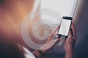 A woman holding and looking at smart phone with blank white screen next to an airplane window with clouds and sky background