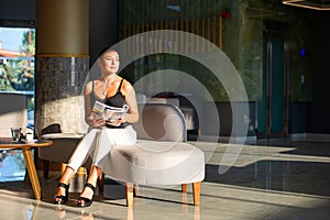 Woman holding magazine, sitting on sofa in light modern interior of hotel reception, office building elevator hall, waiting area