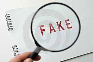 Woman holding magnifying glass over notebook with word Fake on white background, closeup