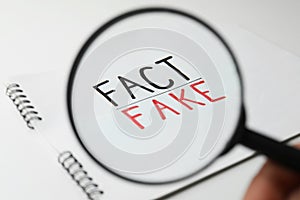 Woman holding magnifying glass over notebook with words Fact and Fake on white background, closeup