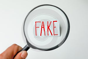 Woman holding magnifying glass over sheet of paper with word Fake on light background, top view