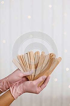 Woman holding many wax wooden spatulas. The young woman is wearing beige clothing and in pink medical gloves. The photo