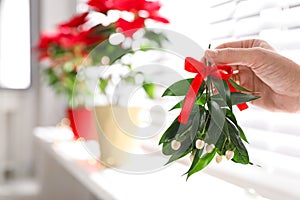 Woman holding mistletoe bunch with red bow indoors, closeup. Traditional Christmas decor