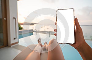 Woman holding mobile phone with blank screen while laying on sunbathing bed with beach view