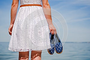 Woman holding moccasins on the beach walking in white dress