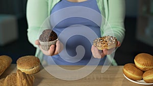 Woman holding muffin and donut, lots of pastry on table, overeating sweets