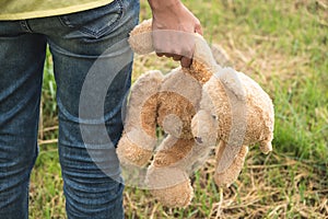 A woman holding an old brown teddy bear with his right hand and walking to the grass field which is a doll from the important day