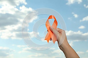Woman holding orange ribbon against blue sky, closeup with space for text. Multiple sclerosis awareness
