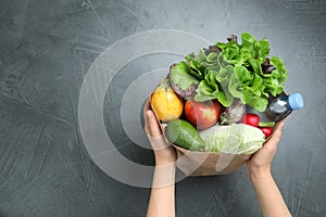 Woman holding paper package full of fresh vegetables and fruits on dark background, top view