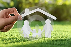 Woman holding paper roof over cutout of family on fresh grass, closeup. Life insurance