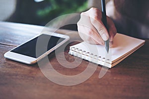 A woman holding pencil and writing on notebook with white mobile phone on wooden table in cafe