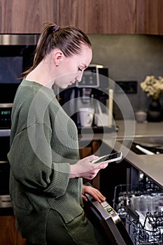 Woman is holding a phone with the smart home program running, remotely controlling the dishwasher. The concept of a