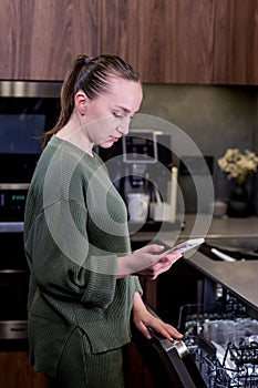 Woman is holding a phone with the smart home program running, remotely controlling the dishwasher. The concept of a