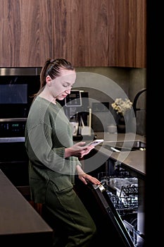 Woman is holding a phone with the smart home program running, remotely controlling the dishwasher. The concept of a