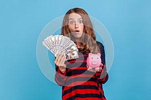 Woman holding piggy bank and big sum of money, looking at camera with surprised expression.