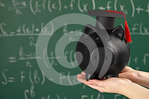 Woman holding piggy bank and graduation cap against greenboard, closeup with space for text. Scholarship concept