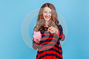 Woman holding piggy bank, pointing to camera with happy expression, offering the way to save money.