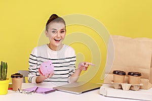 Woman holding piggybank and pointing at coffee with pizza box, looking with positive expression,