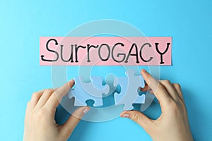 Woman holding puzzle pieces near sheet of paper with word Surrogacy on light blue background, top view