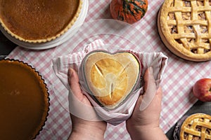Woman holding a raspberry pie, in a heart-shaped tray, above a kitchen table full of traditional fall pies, apple pies, and