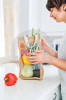 Woman holding reusable eco sackcloth fabric bag packaging with fresh organic vegetables on white kitchen table. Home delivery