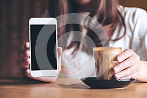 A woman holding and showing white mobile phone with blank black desktop screen with coffee cup on wooden table