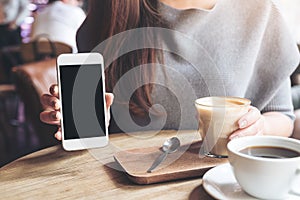 A woman holding and showing white mobile phone with blank black desktop screen with coffee cups on vintage wooden table