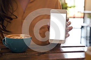 A woman holding and showing white mobile phone with blank desktop screen with coffee cup on wooden table