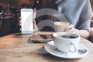 A woman holding and showing white mobile phone with blank desktop screen with coffee cups on vintage wooden table