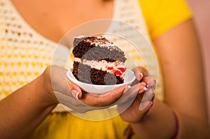 Woman holding small plate of chocolate cake with cream filling, showing to camera, pastry concept