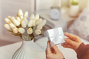 Woman holding small white gift box and greeting card with bouquet of white tulips flowers on valentines day.