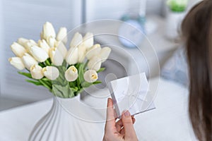 Woman holding small white gift box and greeting card with bouquet of white tulips flowers on valentines day.