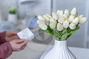 Woman holding small white gift box with jewelry and bouquet of white tulips flowers sitting at the table in home room. Concept.
