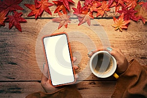 Woman holding smart phone and coffee cup wooden table with autumn maple leaves.