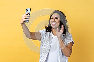 Woman holding smart phone making selfie or making video call, waving hello gesture with hand.