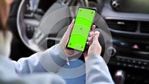 a woman holding a smartphone with a green screen on the background of the steering wheel in the car