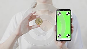 A woman is holding a smartphone with a green screen and a gold bitcoin coin. Close-up. Mock up. The concept of