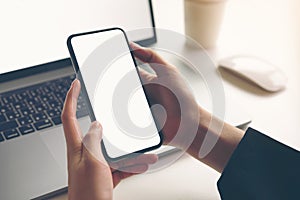 Woman holding smartphone mockup of blank screen and laptop on the table.