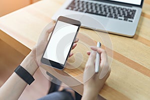 Woman holding smartphone and using laptop on table in office room on windows with trees and nature background.