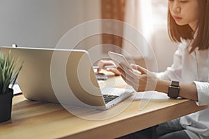 Woman holding smartphone and using laptop on table in office room on windows with trees and nature background.
