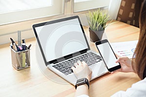 Woman holding smartphone and using laptop on table in office room on windows with trees and nature background.