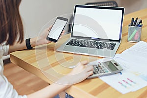 Woman holding smartphone and using laptop on table in office room on windows with trees and nature background.