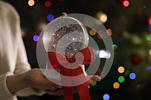 Woman holding snow globe with red bow knot on blurred background, closeup