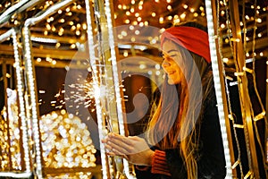 Woman holding sparkler night while celebrating Christmas outside. Dressed in a fur coat and a red headband. Blurred